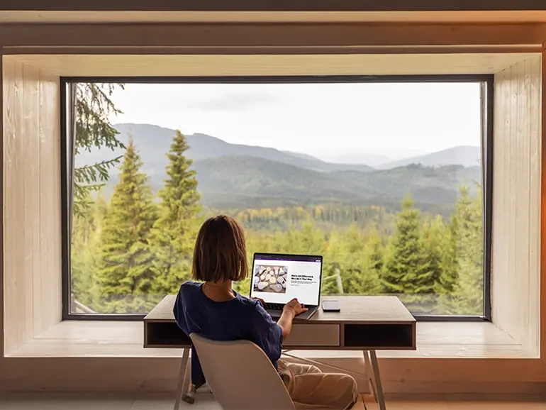 Woman working on laptop in front of window overlooking mountainscape.