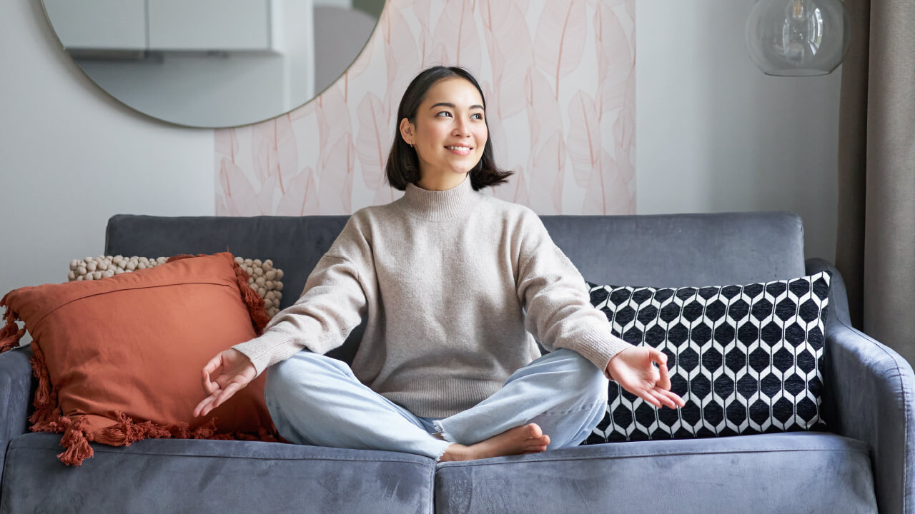 Woman meditating cross-legged on a grey couch. 