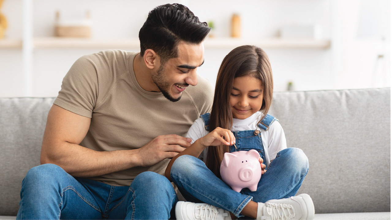 Father and daughter sitting on a couch and putting a coin into a piggy bank. 