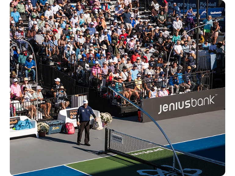 Spectators watch a pickleball match from bleachers on a sunny day. Jenius Bank TM logo is on the side of the court. 