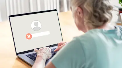 A woman sits at a table and attempts to enter her login to an account that has been taken over by fraudsters.