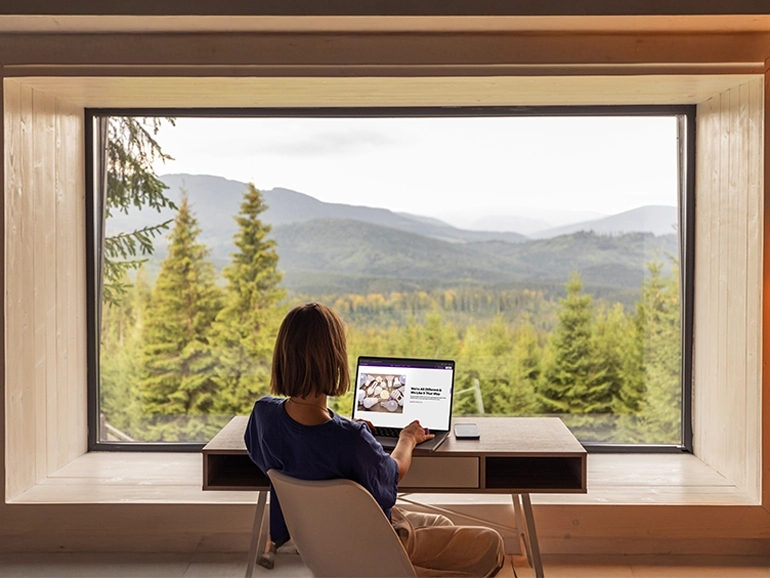 Woman working on laptop in front of window overlooking mountainscape. 