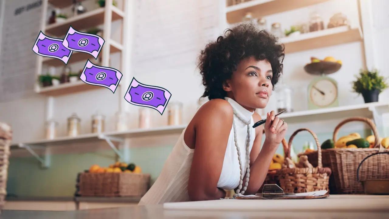 Woman sitting at a kitchen table with a thoughtful expression. Illustrated dollar bills are floating behind her. 